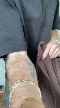 Personne portant un t-shirt noir tenant un vêtement marron, montrant un avant-bras tatoué avec des bracelets en or et en argent.