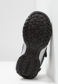 Suela de zapato de goma negra con patrones de agarre circulares y un acento lateral blanco, que presenta un diseño geométrico texturizado y un detalle del logo.