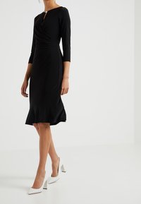 Czarna dopasowana sukienka z rękawami o długości trzy czwarte, warstwową teksturą w talii oraz rozkloszowanym dołem. Zestawiona z białymi butami na obcasie w kształcie bloku.