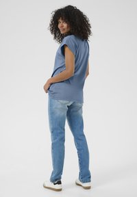 T-shirt blu a maniche corte con bordi arrotolati, abbinato a jeans in denim chiaro e sneakers bianche. Il modello è visto da dietro.