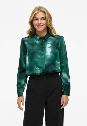 Zelena svilenka sa uzorkom u tehnici tye-dye, dugmad s prednje strane, dugi rukavi i strukturirani ovratnik. Uparena s crnim hlačama.