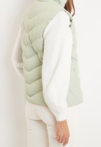 Smanicato imbottito verde chiaro con design trapuntato, abbinato a un maglione bianco a maglia. Senza maniche, con vestibilità rilassata e texture morbida.