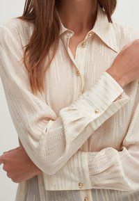 Blusa di chiffon crema con righe verticali dorate, colletto classico e polsini con bottoni. Presenta una sottile texture e raffinati dettagli in metallo dorato.
