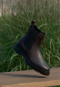 Braune Leder Chelsea-Stiefel mit schwarzen elastischen Seitenpaneelen, runder Spitze und strukturiertem Gummisohle, vor einem verschwommenen grünen Hintergrund präsentiert.