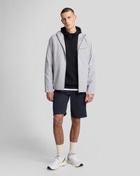 Mladi moški stoji v svetlo sivi kapucarji z zadrgo, črni kapuci, temno modrih kratkih hlačah, belih nogavicah in belih supergah na enobarvnem ozadju.