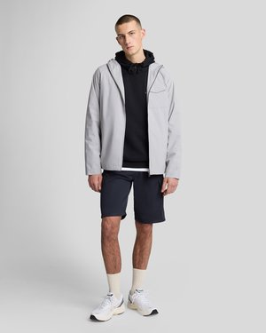 Joven de pie con chaqueta con capucha de color gris claro, sudadera negra, pantalones cortos azul marino, calcetines blancos y zapatillas blancas, frente a un fondo liso.