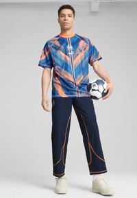 Chemise de sport à motifs bleus et oranges avec des manches courtes, associée à un pantalon marine avec des accents orange, et un ballon de soccer noir et blanc.