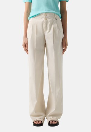 Frau trägt hoch taillierte, weit geschnittene beige Hosen mit zwei Knöpfen und Falten, kombiniert mit schwarzen offenen Sandalen und einem hellblauen Hemd.