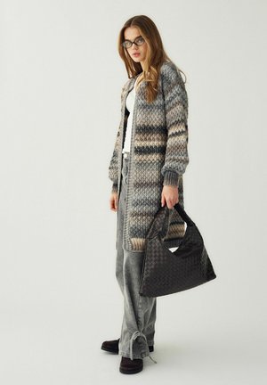 Tânără purtând ochelari, un cardigan lung tricotat cu nuanțe multiple, blugi gri și pantofi negri, ținând o geantă mare neagră țesută.