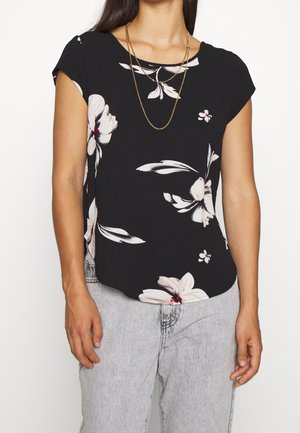 Femme portant un chemisier noir à manches courtes avec un imprimé floral blanc et des colliers dorés superposés, associé à un jean gris clair.