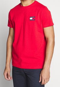 Mężczyzna ubrany w jasnoczerwoną koszulkę Tommy Jeans z małą naszywką na klatce piersiowej i ciemnymi spodniami, stojący na jednolitym tle.