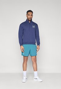 Μπλε πουλόβερ με μισό φερμουάρ και λογότυπο, συνδυασμένο με τιρκουάζ σορτς, λευκές κάλτσες και αθλητικά παπούτσια. Αποπνέει έναν χαλαρό αθλητικό στυλ.