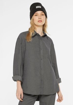 Young woman wearing a dark grey button-up shirt with rolled sleeves, matching pants, and a black beanie labeled "Senses The Label."