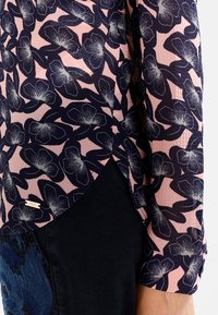 Marinblus med rosa inslag som har ett blommönster, strukturerad tyg, långa ärmar och en något rundad nederkant. Liten varumärkesetikett synlig.