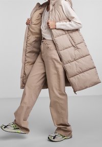 Béžová prošívaná dlouhá vesta s kapucí, nošená přes světlejší svetr, spárovaná s volnými béžovými kalhotami a vícebarevnými sportovními botami.