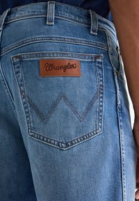 Κοντινό πλάνο πίσω τσέπης μπλε τζιν με διπλό ραφτό μοτίβο "W" και καφέ δερμάτινη ετικέτα της μάρκας Wrangler πάνω από την τσέπη.