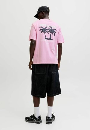 Osoba ubrana w różowy T-shirt z czarną grafiką palmy, czarne spodenki do kolan, białe skarpetki, czarne buty sportowe i czapkę z daszkiem, zwrócona tyłem.