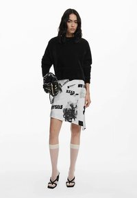 Juodas pūkuotas megztinis, asimetriška balta sijonas su juodu grafiniu tekstu, permatomos šviesios kojinės ir juodos basutės su dirželiais. Papildyta raštuota rankine.