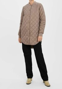 Pikowana długa kurtka w jasnobrązowym kolorze, z zapięciem na guziki, stojącym kołnierzem i fakturą w kształcie diamentów. Do tego czarne spodnie i paskowane szpilki.