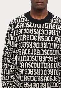 Maglione lavorato a maglia bianco e nero con ripetuti design grafici in testo. Il collo a girocollo e i polsini a coste ne esaltano l'aspetto texturizzato.