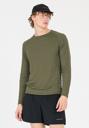 Jonge man met een zwarte pet, een olijfgroen shirt met lange mouwen en zwarte shorts, staand tegen een effen witte achtergrond.