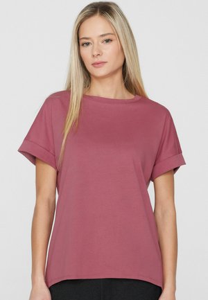 Femme blonde aux cheveux raides portant un T-shirt mauve à manches courtes retroussées, posant devant un fond blanc uni.