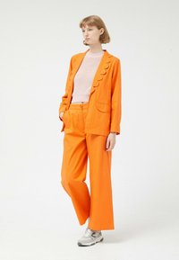 Blazer naranja con solapas en forma de concha, combinado con pantalones de pierna ancha a juego. La modelo lleva una camiseta texturizada de color rosa claro y zapatillas grises.