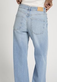 Osoba u svijetloplavim trapericama visokog struka s džepovima na leđima i tamnobraon kožnim natpisom marke, prilagođava pojas rukama.