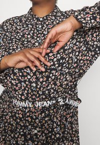 Vestido negro con flores pequeñas y coloridas. Presenta un cuello abotonado y una cinturilla blanca con el logo "TOMMY JEANS". Mangas largas.