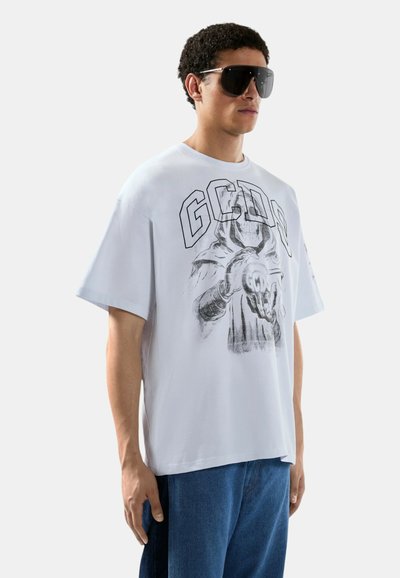 Homme portant un T-shirt blanc oversize avec un graphisme représentant une silhouette squelettique tenant une boule lumineuse et un grand texte « GCDS », associé à un jean bleu et des lunettes de soleil.