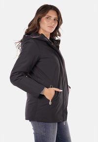Giacca nera impermeabile con cappuccio, tasche con zip, vita aderente e texture liscia. Progettata per calore e comfort.