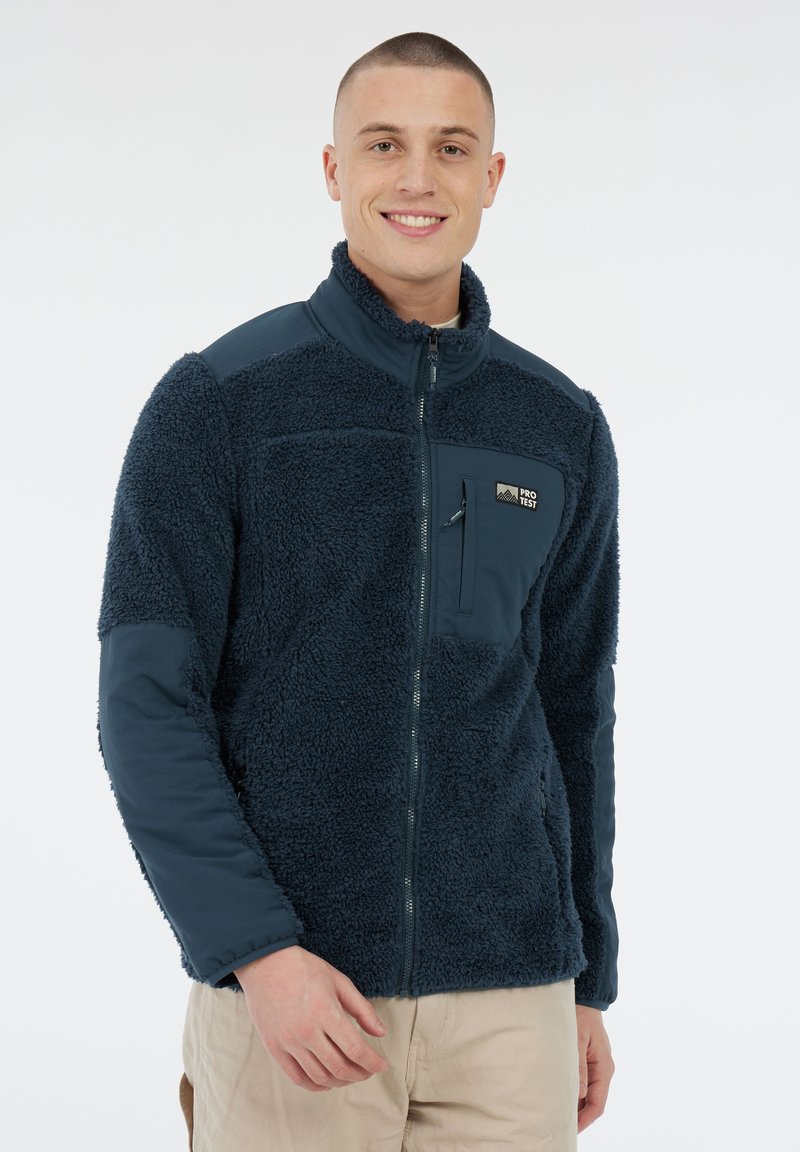 Glunderende man in een marineblauwe fleecejack met rits en borstzak, staande tegen een effen witte achtergrond.