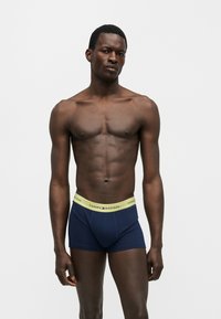 Άνδρας μοντέλο με μυώδες σώμα που φοράει σκούρα μπλε μπόξερ με κίτρινη μέση με την επωνυμία "Tommy Hilfiger" σε απλό λευκό φόντο.
