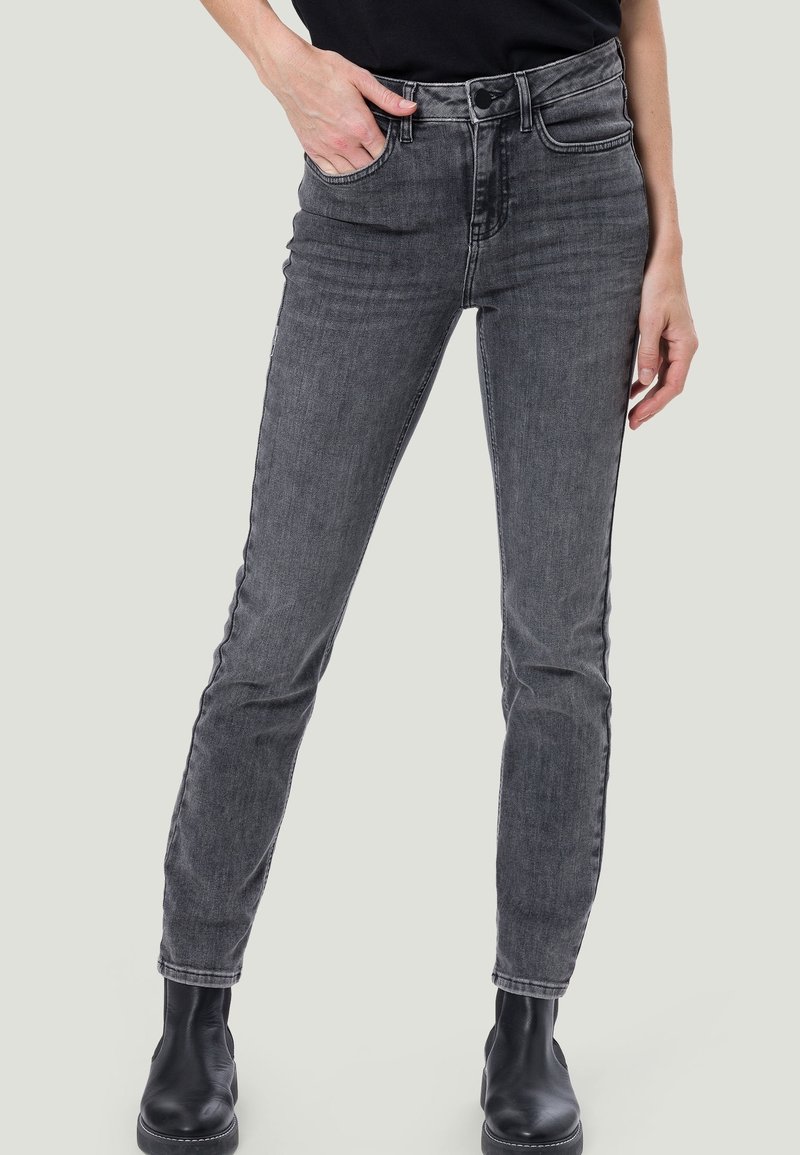 Jean gris slim fit porté avec des bottes noires et un haut noir, une main dans la poche et l'autre détendue sur le côté.
