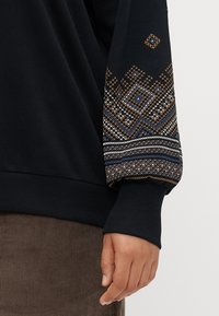 Μαύρη φούτερ μπλούζα με διακοσμημένα μανίκια κεντημένα που διαθέτουν γεωμετρικά σχέδια σε μπλε, λευκό και χρυσό. Ριμπ μανσέτες και τελείωμα.