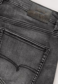 Gros plan sur un jean denim gris foncé montrant la poche arrière et une étiquette en cuir noir avec le texte "DENIM SUPPLY".