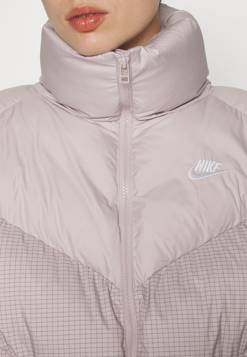 Veste matelassée violet clair avec col montant, design en zigzag, section inférieure à carreaux et logo Nike blanc sur la poitrine. Fermeture éclair à l'avant.