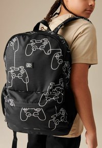Schwarzer Rucksack mit einem Muster aus weißen Videospielcontrollern, aus strapazierfähigem Stoff mit einem Reißverschluss und verstellbaren Trägern.