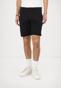 Shorts noirs à coupe classique, fabriqués en tissu doux. Présentent un bouton de fermeture, un ourlet retourné et des poches avant. Portés avec des baskets blanches.