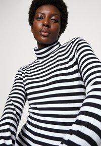 Black and white striped turtleneck sweater with ribbed texture. Features a small logo detail on the chest and fitted long sleeves.