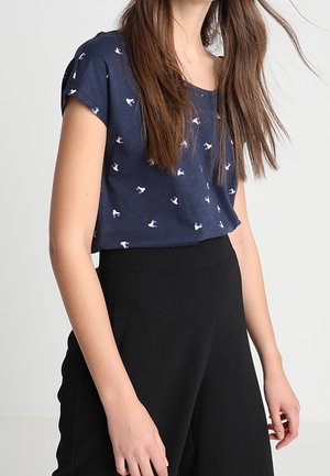 Kobieta ubrana w granatową koszulkę z krótkim rękawem z białymi nadrukami ptaków oraz czarne spodnie z wysokim stanem, stojąca na jednolitym tle.