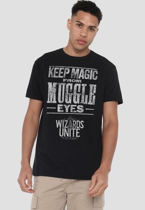 Young man wearing a black T-shirt with white text reading "Keep magic from muggle eyes" and "Wizards Unite," beige cargo shorts.