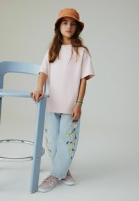 Maglietta rosa chiaro oversize, jeans blu ricamati con motivi floreali e sneaker rosa. Il bambino indossa un cappello a tesa larga marrone e sta accanto a una sedia blu.