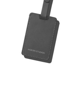 Étiquette de bagages en cuir noir avec des bords cousus et une boucle pour l'attache. Présente le logo en relief "HORIZN STUDIOS" en bas.