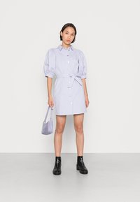 Vestido de botão lilás claro com mangas curtas e volumosas e um cinto na cintura. Combinado com uma pequena bolsa texturizada roxa e botas de tornozelo pretas.