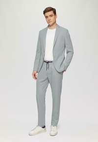 Hellgrauer Anzug mit taillierter Passform, bestehend aus einem einreihigen Blazer und passender Hose. Tragen über einem weißen T-Shirt, kombiniert mit weißen Sneakers.