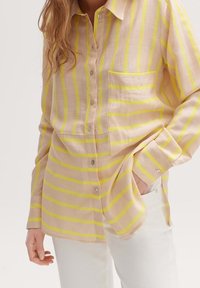 Long-sleeve, button-up shirt in light tan with bold yellow stripes. Features a chest pocket and rounded hemline. Made of lightweight fabric.