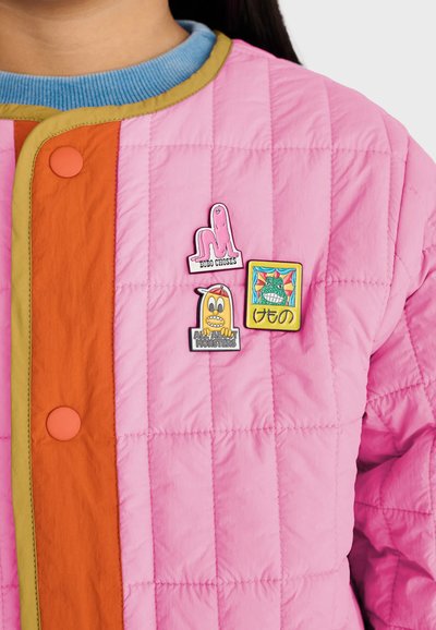 Personne portant une veste matelassée rose avec des boutons orange et trois badges colorés de dessins animés sur la zone de la poitrine.