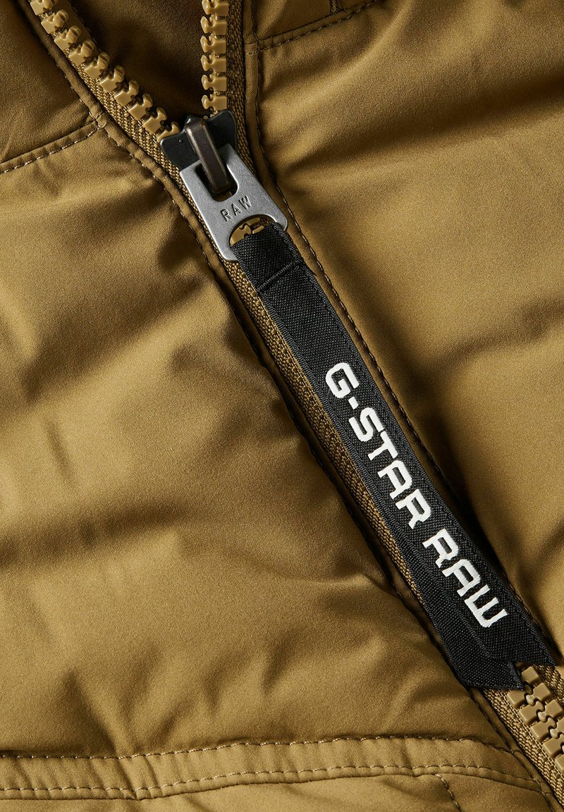 Zbliżenie na beżową kurtkę z czarnym uchwytem zamka błyskawicznego, oznaczoną etykietą "G-STAR RAW", z metalowym suwakiem zamka z wygrawerowanym napisem "RAW".