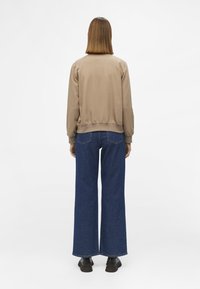 Beige bomberjack met elastische manchetten en tailleband, gecombineerd met hoge, wijde blauwe jeans en zwarte schoenen. Eenvoudig, strak ontwerp.